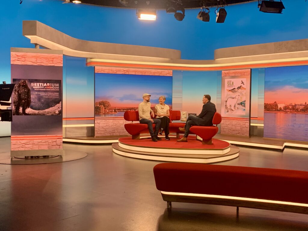 Fernsehstudio mit Gästen und Moderator. Sven Nieder, Laali Lyberth und Holger Wienphal. Links: Bestiarium Groenlandicum. Rechts: Sila. 